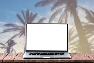 Modern laptop with empty white screen on wooden table against blurred palm trees background