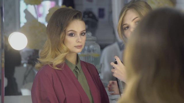 Stylist corrects lacquer styling on voluminous hair of female model. In front of mirror there is woman, whom a hairdresser is making fashionable hairstyle in beauty salon.