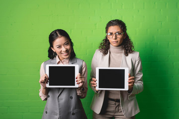 Fototapeta na wymiar african american and asian businesswomen showing digital tablets in front of green wall
