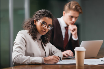 african american businesswoman writing in documents while businessman using laptop