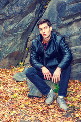 American man autumn/winter casual street fashion. Young handsome man wearing black leather jacket, pants, gray leather shoes, sits on rocks with fallen leaves on ground in Central Park, New York..