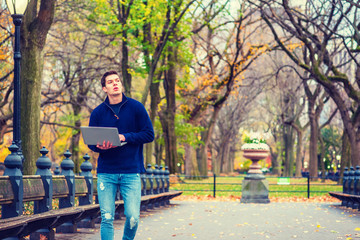 Way to Success. American Man wearing blue sweatshirt, fashionable jeans, walks on road with long bench, trees at Central Park, New York in autumn day, reads, works on laptop computer. Filtered effect.