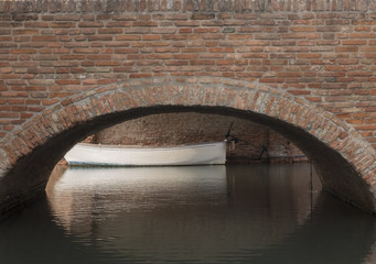 Brick bridge in sight and its arch reflecting on the underlying canal of Comacchio, Italy. Small white boat moored just behind the bridge