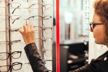 Pretty young woman is choosing new glasses at optics store.
