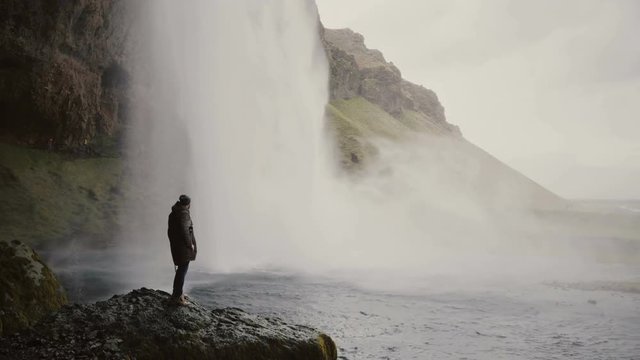 Young man with action camera standing close to Gljufrabui waterfall in Iceland and filming video of stream of water.