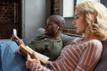 african american man listening music while caucasian girlfriend reading book on sofa at home