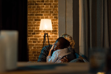 selective focus of multiethnic couple hugging each other while sitting on sofa at home