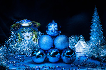 Pile of blue christmas balls in the center surrounded with silver tinsel. Black and blue background. Carnival mask, xmas tree and house behind.