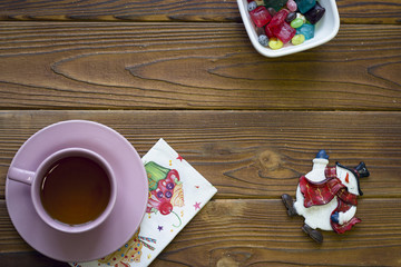 Pink tea cup with saucer, toy snowman and a bowl of colored candies on a rustic wooden background, top view