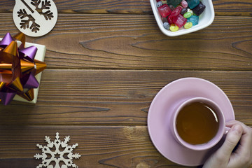 Hand holding a pink tea cup, gift box, wooden toys and a bowl of colored candies on a rustic wooden background, top view