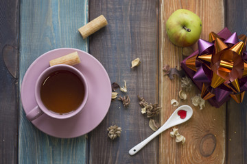 A tea cup,porcelian spoon,gift box with a shiny bow and an apple on a colored wooden table, top view