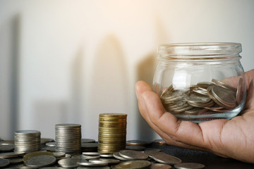 Hand holding jar saving money with stack of coins