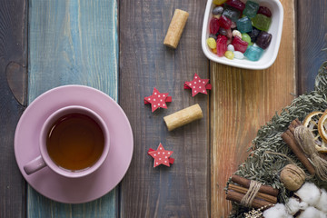 Christmas still life with tea cup,saucer, pine wreath and a bowl of colored candies and jelly beans on a colored wooden background, top view