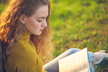 Beautiful young blonde woman sitting and reading book in a park near the lake