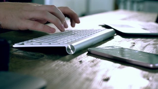 Closeup on a caucasian male freelance designer, programer working at Home typing on a wireless keyboard with electronics peripheral on the wooden table
