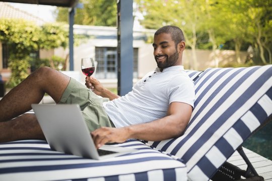 Man holding wineglass while using laptop on lounge chair