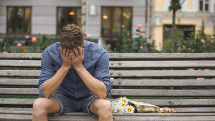 Upset young man sitting on bench alone, closing face with hands, break-up