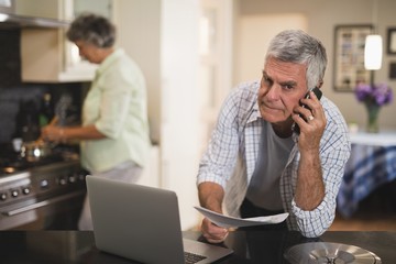 Portrait of senior man talking on phone while wife cooking in