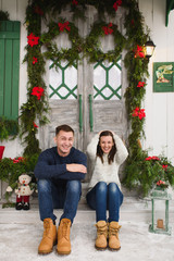 Happy funny young couple in love dressed in sweater sitting on porch steps at light house with decorated in red green New Year door at home. Christmas good mood. Family and holiday 2018 concept