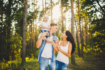 stylish young Family of mom, dad and daughter one year old blonde sitting near father on shoulders, outdoors outside the city in a park amid tall trees in summer at sunset.