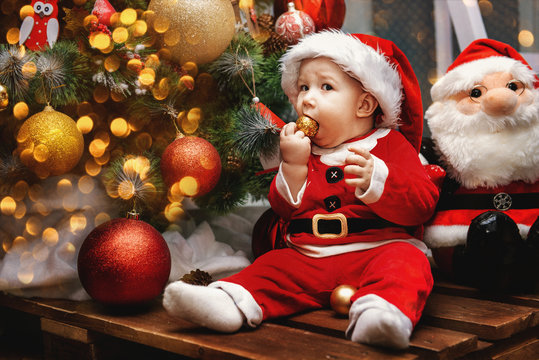 Portrait Caucasian baby boy with gray eyes in red Santa Claus costume sitting by New Year tree, lifestyle Christmas holiday concept