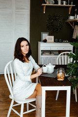 The young woman the brunette sits in kitchen with green walls in the apartment and has breakfast. On a table a cup of tea and cookies. At a background the Christmas fir-tree. 