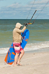 Young male walking, tropical, sandy beach carrying a kiteboard, holding the handlebar of a cabled...