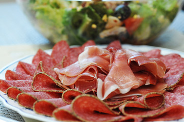 Meat plate with sliced salami and jamon with a bowl of salad on the background