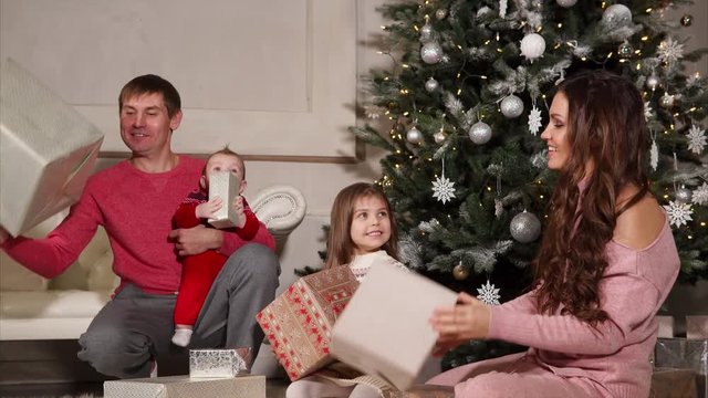 Happy mother and father with kids are taking apart christmas gifts. They are sitting on a floor in a big light hall of home with decorated large spruce.