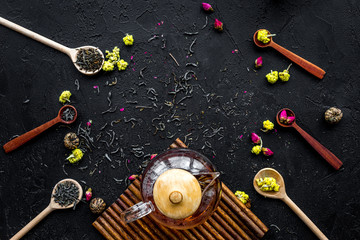 Brew the aromatic tea. Tea pot near wooden spoons with dried tea leaves, flowers and spices on black wooden background top view copyspace