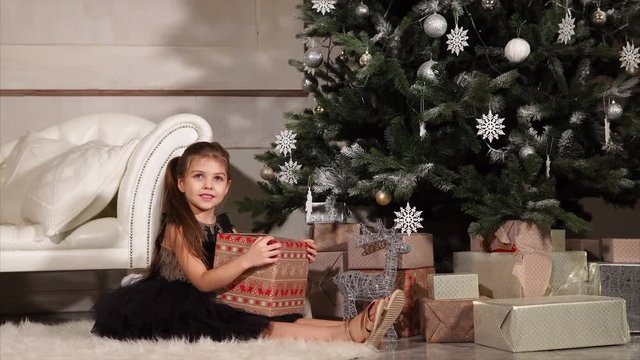 Child girl is well-dressed and happy, is sitting on a carpet in a hall near a big decorated spruce. She is holding large box with Christmas gift and looking on it.