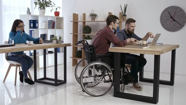 Skillful disabled man in wheelchair discussing new startup project on laptop pc with coworker while asian female colleague talking on phone in creative office. Handicapped freelancer working in office