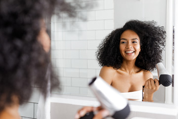 Smiling woman with hair dryer looking at mirror - obrazy, fototapety, plakaty