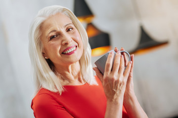 Cup of tea. Nice elderly positive woman holding a cup of tea and smiling to you while resting at...