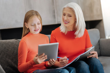 Modern generation. Pleasant cheerful young girl holding a tablet and smiling while sitting together with her grandmother