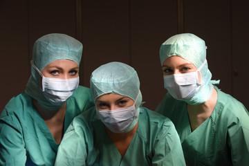 A group of three young women pose in a medical operation theater.  Fully dressed as theater nurses with face masks  and green sterile medical work clothing.
