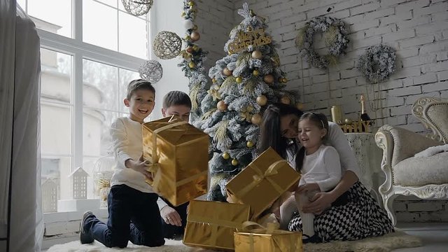 Young family dressed in white clothes, sit on the floor near the Christmas tree in the living room with a large window and rejoice in the gifts that are in golden boxes