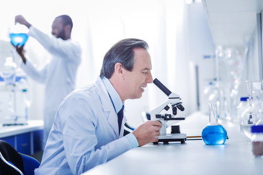 Favourite occupation. Happy nice intelligent scientist looking into the microscope and smiling while enjoying his job