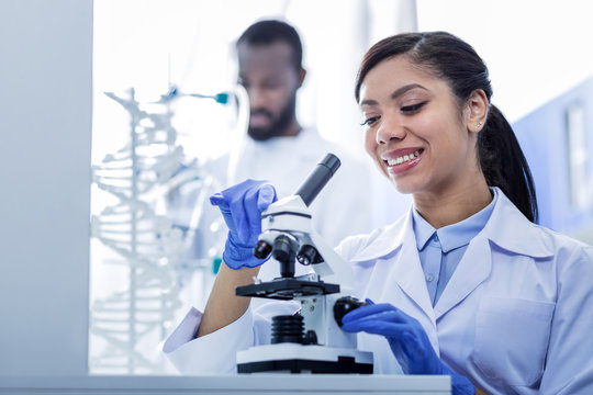 Wonderful mood. Nice positive young woman smiling and looking at the microscope while being excited about the research