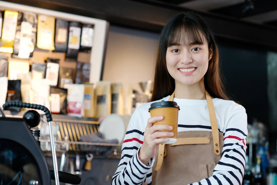 Young asia woman barista holding a diaposable coffee cup with smiling face at cafe counter background, small business owner, food and drink industry