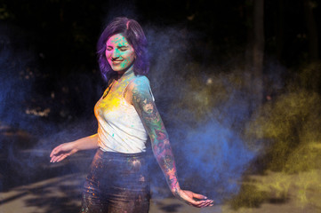 Merry woman with blue and yellow Holi powder exploding around her. Woman posing at the park in sun light