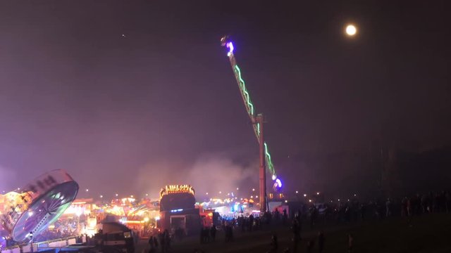 Busy night at traveling travelling carnival, or funfair outdoors under the light of full moon. Many people and children riding colorful illuminated amusement park rides
