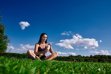 Girl sits in Park on a background of clear sky and opens the water bottle.