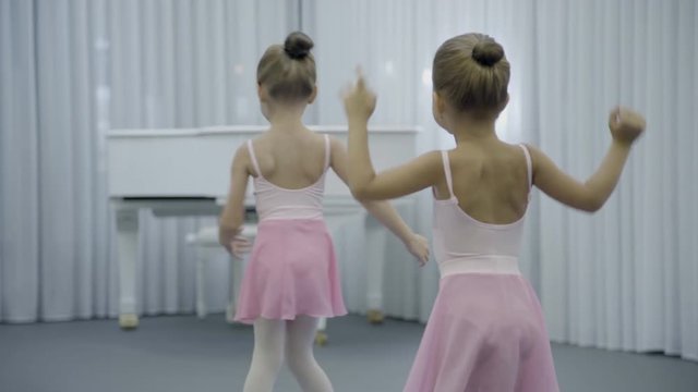 Two girls are having fun in ballet studio indoor. They talk friendly and turn bodies, raising their hands up. Adorable children dressed in classical dance clothes spend good time, learn basics of
