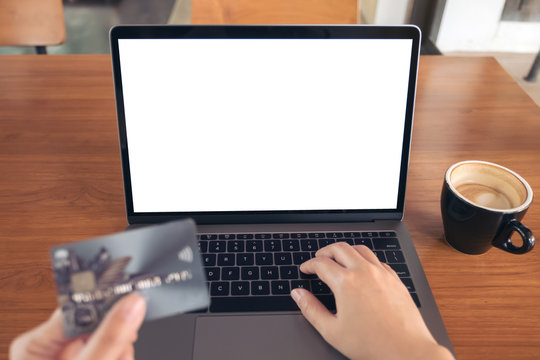 Mockup image of a hand holding credit card while using and typing on laptop with blank white screen and coffee cup on wooden table in modern loft cafe