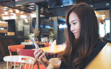 Closeup image of a beautiful Asian woman with smiley face using and looking at smart phone in modern cafe