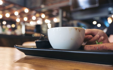 Closeup image of a woman holding a white cup of coffee on black saucer on wooden table in modern cafe