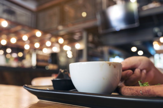 Closeup image of a woman holding a white cup of coffee on black saucer on wooden table in modern cafe