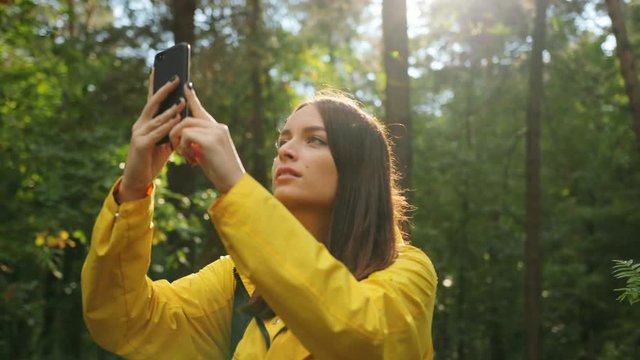 Portrait shot of young pretty woman taking photos of the nature on the black smartphone while hiking in the forest. Sunny day. Outside
