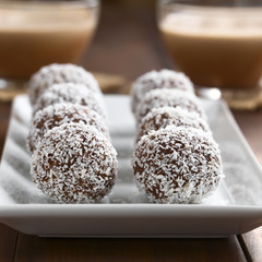 Homemade coconut rum balls on plate, glass cups of hot chocolate in the back, photographed with...
